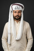A man wearing a traditional white keffiyeh headscarf with black Arabic calligraphy and red decorative trim, paired with a cream-colored long-sleeve shirt, set against a solid dark gray background.